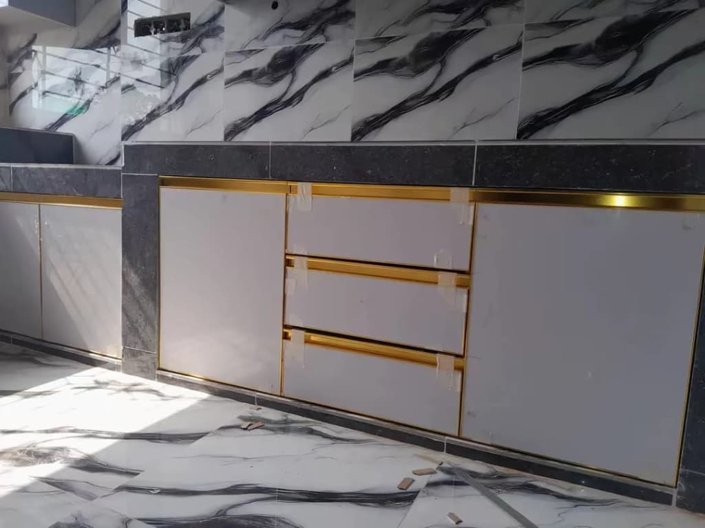 White kitchen drawers lined in gold, set against a dark stone worktop and a marble floor.